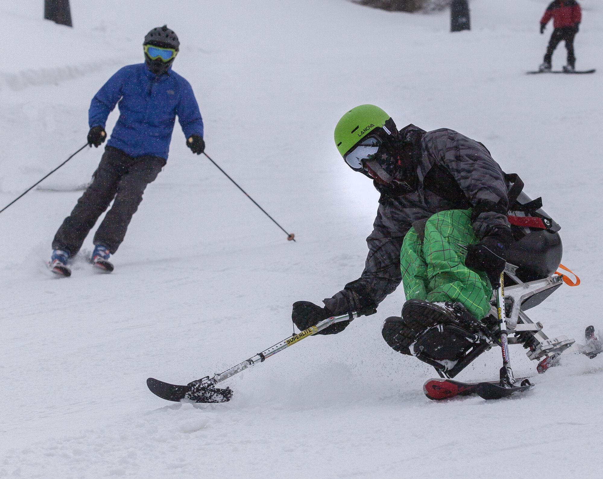 A participant in a green helmet carves a turn in a mono-ski at Big Bear while a USARC volunteer skis close behind.