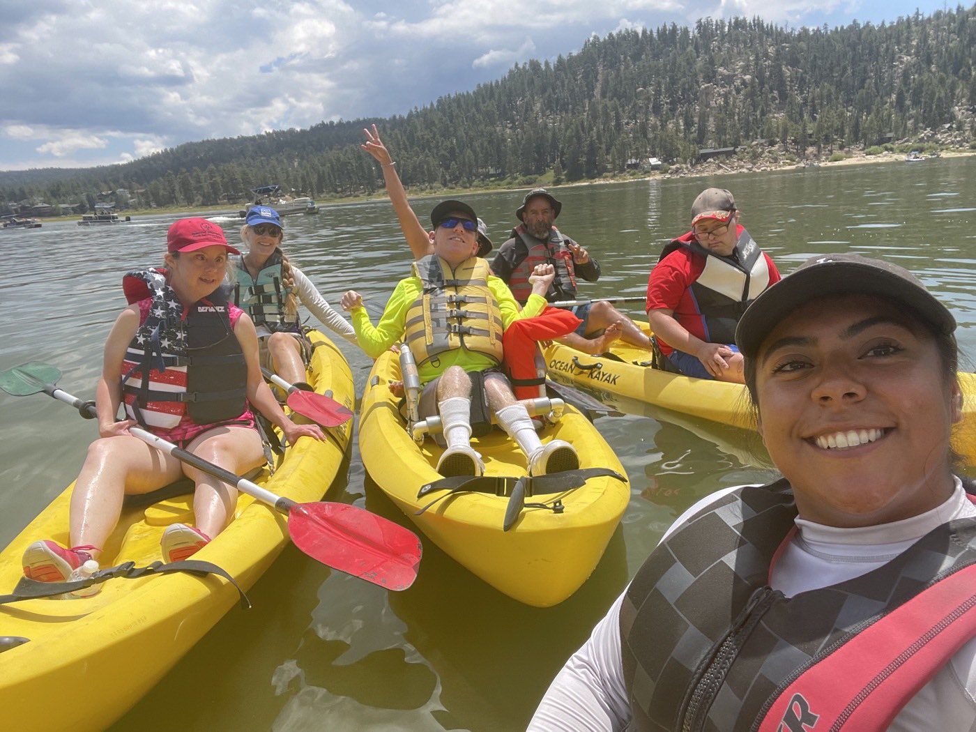 Participants and volunteers in yellow kayaks wave and smile on Big Bear Lake, pine-covered hills behind them.