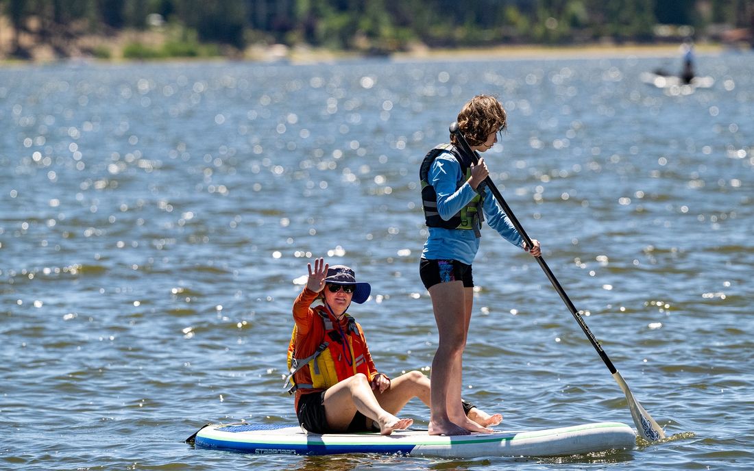 Two paddlers share a stand-up paddleboard on Big Bear Lake, one seated and waving.