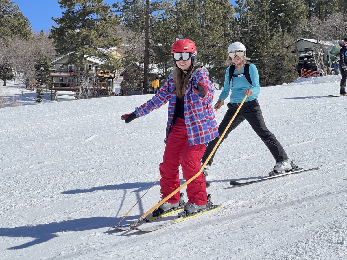 A USARC volunteer skis behind a participant in a red helmet, holding tethers to control speed on a groomed run.