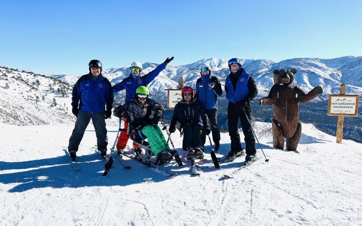 USARC volunteers and participants, one in a sit-ski, celebrate at the top of a run with a bear mascot and snowy peaks behind them.